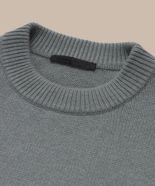 Close-up of the Cashmere Oversized Crewneck in grey, featuring a ribbed crew neckline and black fabric label inside the collar. Displayed against a beige background, this sweater showcases its relaxed pullover style.