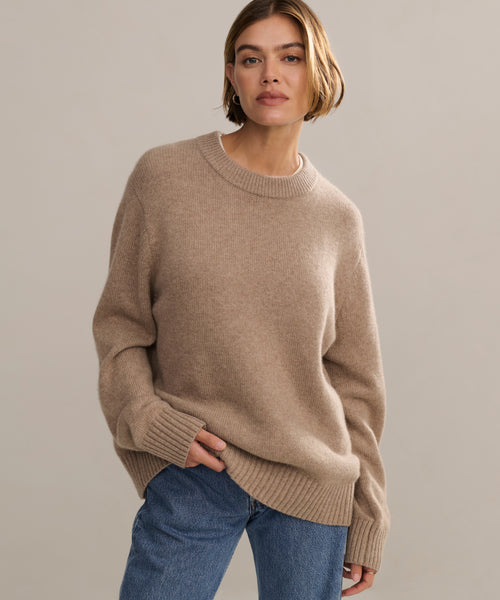 A woman with short blonde hair models the Cashmere Oversized Crewneck with blue jeans, standing against a plain light background, one hand in her pocket.