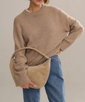 A person wearing the Cashmere Oversized Crewneck in beige and blue jeans holds a large beige suede shoulder bag with a twisted handle. Their head is partially out of frame.