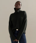 A person models the Cashmere Porter Turtleneck with blue jeans, standing against a beige background and looking confidently at the camera with hands clasped.