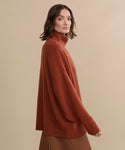 A woman with shoulder-length brown hair wears the Cashmere Porter Turtleneck and a brown skirt, standing against a plain beige background. She looks at the camera with a neutral expression.