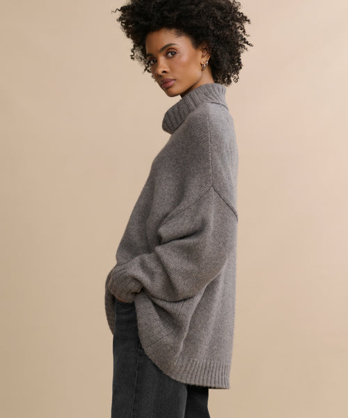 A woman with curly hair wears the oversized Cashmere Porter Turtleneck and dark pants, standing sideways with her hands in her pockets against a plain beige background.