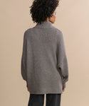 A person with curly hair is seen from the back, wearing the Cashmere Porter Turtleneck—an oversized, loose turtleneck sweater—paired with dark pants, standing against a plain beige background.