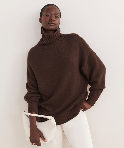 Wearing the Cashmere Porter Turtleneck and white pants, a person leans against a light wall, holding a white handbag. The look is minimal and elegant.
