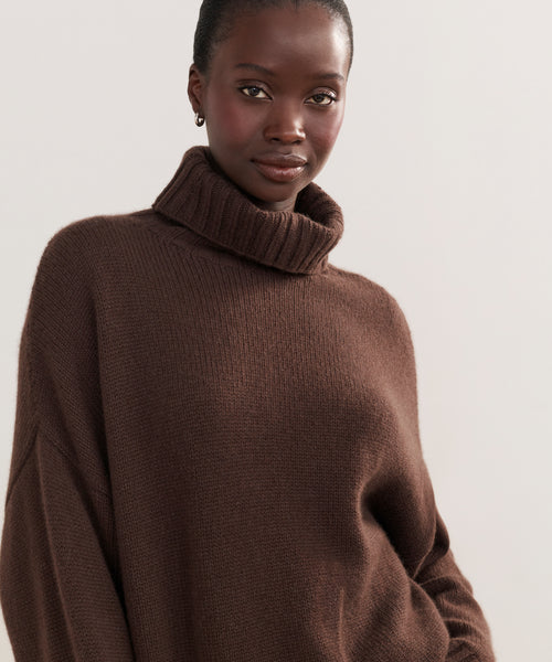 A woman with dark skin and pulled-back hair models the Cashmere Porter Turtleneck, an oversized sweater, while standing confidently against a plain light background.