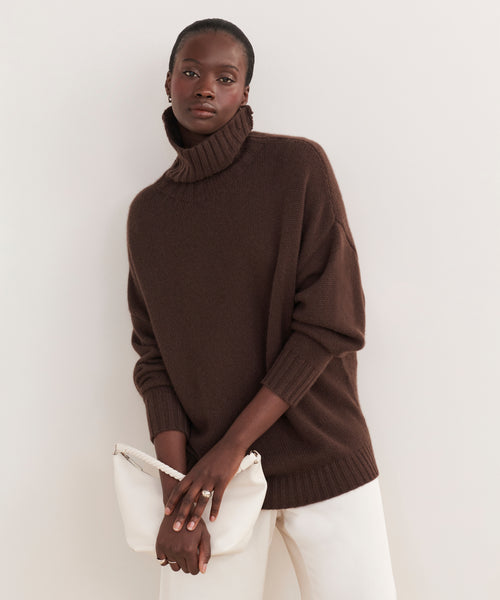 Wearing the Cashmere Porter Turtleneck in dark brown and white pants, a person stands against a light background, holding a white clutch bag with both hands.