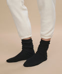 A person wears off-white sweatpants with ankles slightly rolled up, highlighting luxurious Cashmere Socks in black. Standing on a neutral beige background, the socks are perfectly showcased—making them an ideal gift choice.