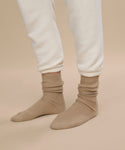 Model showcases caramel beige Cashmere Socks paired with off-white jogger pants against a beige floor and background, highlighting only the lower legs and feet for a cozy autumn/winter mood.