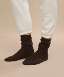 A person wears dark brown Cashmere Socks and cream sweatpants, standing on a beige background with only their lower legs and feet visible—a perfect cozy fall winter gift.