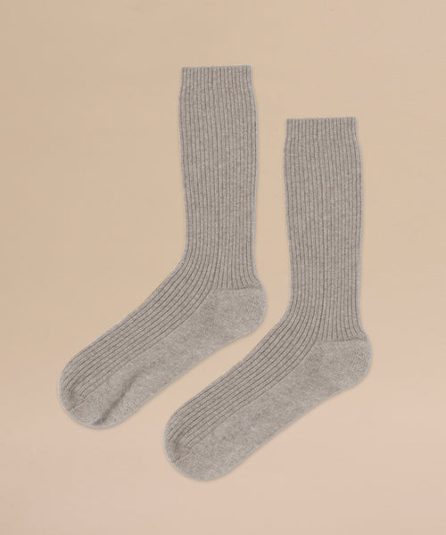 A pair of Cashmere Socks in light gray, ribbed crew style, is laid flat side by side on a beige background.