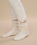 A person wears ivory Cashmere Socks, ideal for fall and winter, paired with off-white sweatpants. Only the lower legs and feet are visible against a light beige surface and neutral beige background.