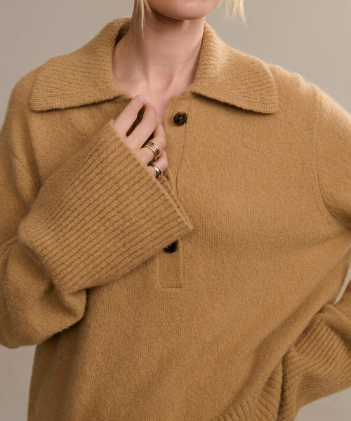 A person wears the Cashmere Stevie Polo, a camel knit top with large ribbed sleeves and an oversized collar, holding the sweater near the collar to display several gold rings. The persons face is partially out of frame.
