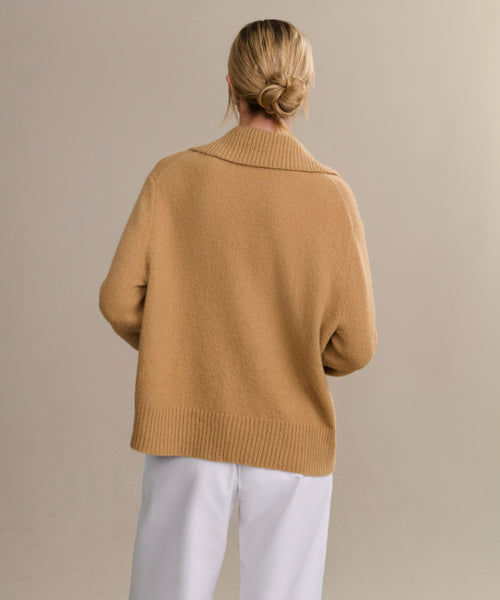 A person with blonde hair in a low bun wears the Cashmere Stevie Polo sweater and white pants, standing with their back to the camera against a plain beige background.