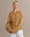 A woman with blonde hair in a low bun wears the Cashmere Stevie Polo in camel and white pants, holding a matching tan handbag against a plain beige background.