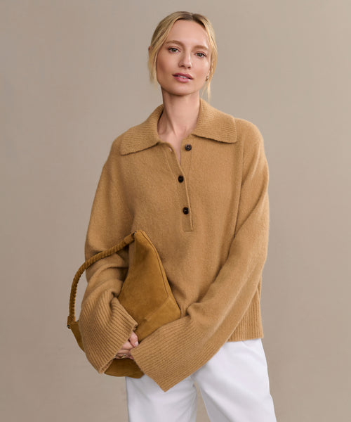 A woman with blonde hair in a low bun wears the Cashmere Stevie Polo in camel and white pants, holding a matching tan handbag against a plain beige background.