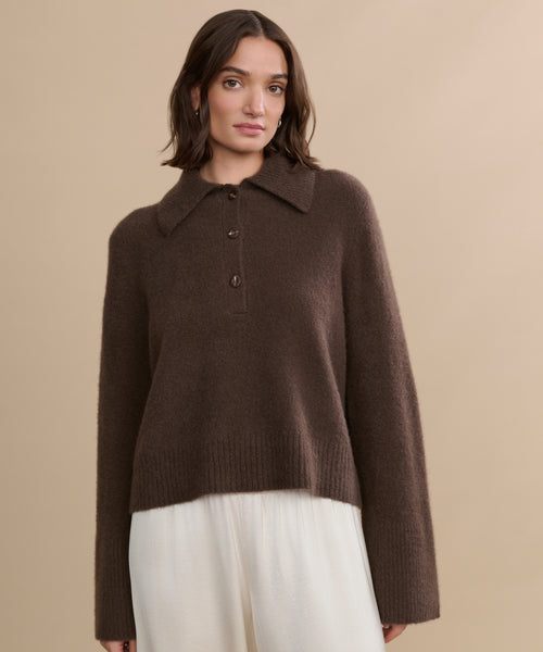 A woman with medium-length brown hair wears the Cashmere Stevie Polo, featuring an oversized collar and buttons, paired with loose white pants against a beige background.