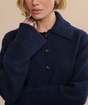 A woman with a short blonde bob wears the Cashmere Stevie Polo in navy, featuring long sleeves and a ribbed collar. She displays rings on her hand near her face against a beige background.