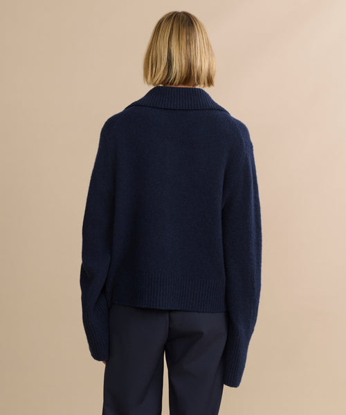 A person with short blonde hair, seen from behind, wears the Cashmere Stevie Polo in navy with an oversized collar and dark pants, standing against a plain beige background.