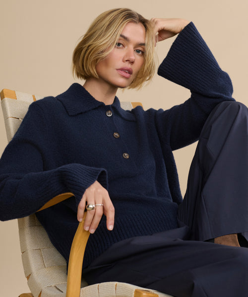 A person with short blonde hair sits in a wooden chair, wearing the navy Cashmere Stevie Polo with large sleeves and dark pants, resting one arm on the chair’s back and the other on their raised knee, set against a beige background.