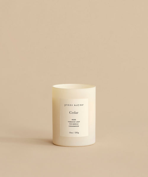 The Cedar Glass Candle, hand-poured in Los Angeles, features a minimalist white design and scent notes of rose, tobacco leaf, and cedarwood. It is shown on a beige surface with a matching beige background.