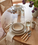 A wooden dining table set with neutral-toned plates, bowls, mugs, the Bistro Vintage Finish Cutlery Set with white handles, glasses of water, a white napkin, and a vase of purple tulips. Sunlight streams onto the table.