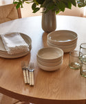 A wooden table set with stacked beige stoneware plates and Pacific Soup Bowls, four metal forks with white handles, three clear glasses, a white napkin, and a vase of green leafy branches.