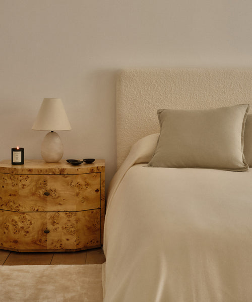 A minimalist bedroom with a cream-colored bed featuring the Chateau Alpaca Coverlet, a light gray pillow, and a wooden nightstand holding a lamp, lit candle, and two small bowls against a plain light wall.