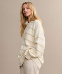 A woman with long blonde hair models the Chloe Cardigan, a loose cream knit with light beige stripes, paired with light pants—an effortless spring and summer staple. She poses with one hand in her pocket against a beige background.