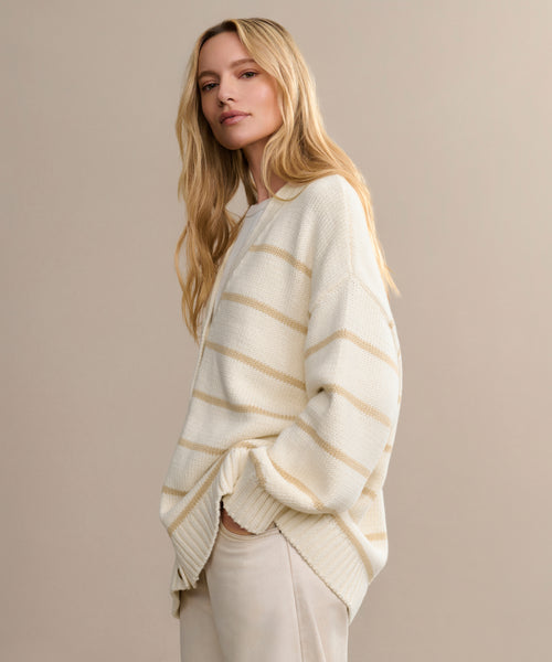 A woman with long blonde hair models the Chloe Cardigan, a loose cream knit with light beige stripes, paired with light pants—an effortless spring and summer staple. She poses with one hand in her pocket against a beige background.