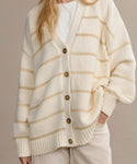 Wearing the Chloe Cardigan—an oversized cream sweater with tan stripes and brown buttons—over a white shirt and beige pants creates a perfect spring/summer look. This warm-weather cardigan features ribbed cuffs and hem. Head out of frame.