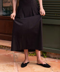 A person in a black midi skirt and Clara Mule shoes stands on a tiled floor, holding their skirt with both hands. Their upper body and face are out of frame.