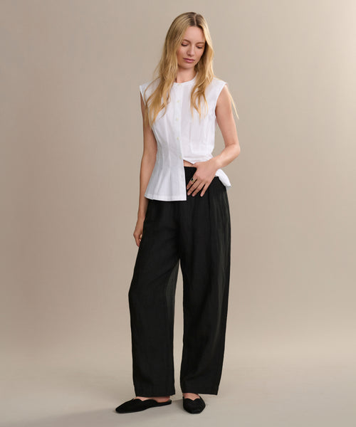 A woman with long blonde hair wears a sleeveless white blouse, loose black pants, and the Clara Mule shoes. She stands against a beige background, looking slightly down with one hand in her pocket.