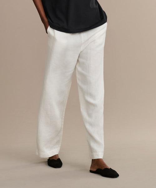 A person in loose white linen pants, a black shirt, and Clara Mule black mules stands against a beige background with hands in pockets; their face is not visible.