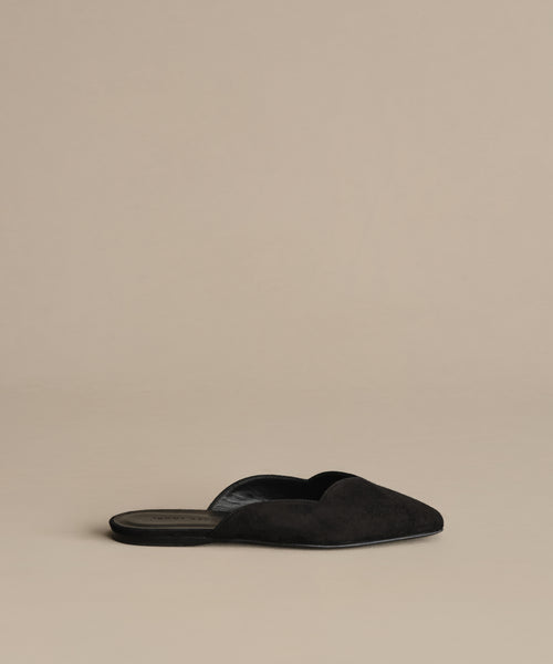 A single Clara Mule in black, featuring a pointed toe and suede finish, is elegantly displayed on a neutral beige background.