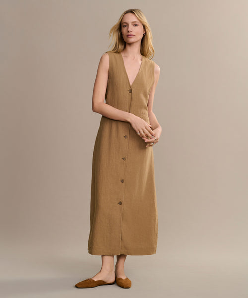 A woman with light hair stands against a beige background, wearing a sleeveless tan dress with front buttons and the Clara Mule shoes in cognac. She appears relaxed, showcasing the Italian-made Clara Mule.
