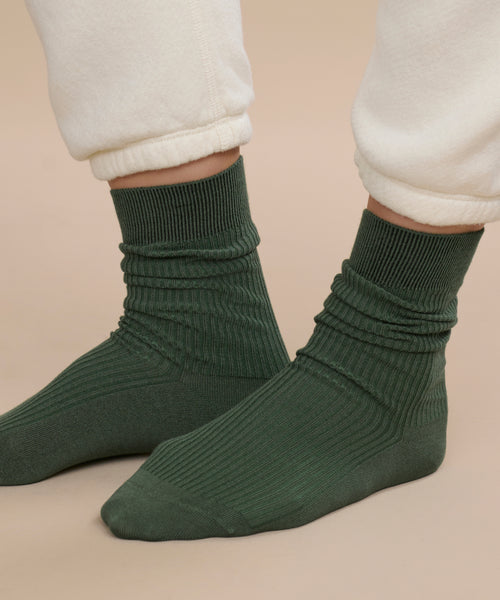 A person wearing cream-colored cuffed pants and Classic Socks in dark green with a ribbed, breathable design, standing on a beige background.