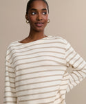 A woman with dark skin and hoop earrings smiles softly. She wears the Claude Long-Sleeve Tee, made of 100% cotton, featuring light beige nautical stripes on a neutral background.