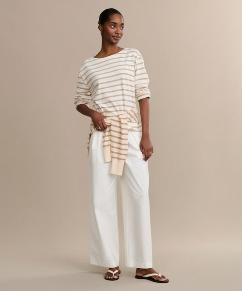 A woman stands against a beige background wearing the Claude Long-Sleeve Tee in cream and tan stripes, paired with white wide-leg pants, sandals, and a matching striped sweater tied at her waist.