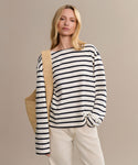 A blonde woman wears the Claude Long-Sleeve Tee with nautical stripes and 100% cotton, paired with light beige pants and a woven straw bag, standing against a plain beige background.