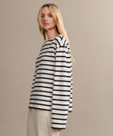 A woman with long blonde hair wears the Claude Long-Sleeve Tee—an oversized, 100% cotton top with black nautical stripes—and loose beige pants, standing against a plain beige background.