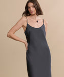 A woman with wavy brown hair models the sleeveless Cleo Slip Dress in dark gray, featuring thin straps, accessorized with a black pendant necklace. She stands against a plain beige background with one hand on her hip.