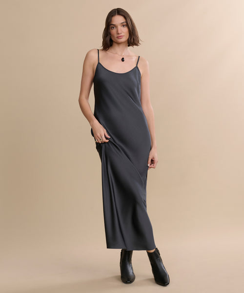 A woman with short brown hair wears the Cleo Slip Dress, a sleeveless dark gray satin style, paired with black ankle boots, standing relaxed against a beige background.