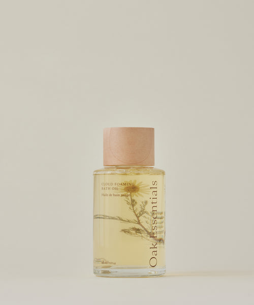 The Cloud Foaming Bath Oil comes in a clear bottle with a light wood cap, holding pale yellow oil and a dried plant sprig inside—creating a spa-like bath experience set against a neutral, light background.