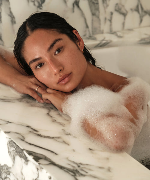 A person with wet hair rests their chin on folded arms at the edge of a marble tub, surrounded by bubbles from Cloud Foaming Bath Oil, gazing calmly at the camera.