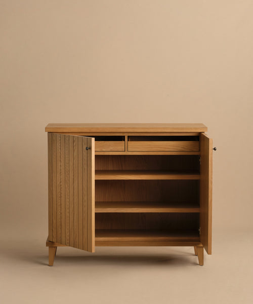 The Coast Hutch is a stylish wooden cabinet with two open doors, two shelves, and two small drawers. Its angled legs and light wood finish bring mid-century modern charm to any space, shown here against a beige background.