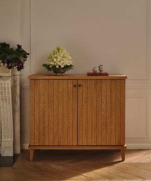 The Coast Hutch, crafted from solid white oak, stands against a beige wall with a glass vase of white flowers and a red tray holding two teacups on top. Next to it is a white marble fireplace adorned with floral arrangements.