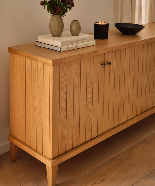 The Coast Sideboard with vertical slats stands on a wooden floor, topped with stacked books, a candle, a black bowl, and a vase of flowers. Sunlight streams in from a window with beige curtains nearby.
