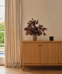 A Coast Sideboard stands against a light wall, topped with a vase of dark red foliage and a lit black candle. Cream-colored curtains frame the window to the left, showing greenery outside.