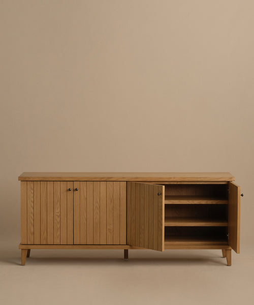 The Coast Sideboard in warm oak has four doors—three closed and one open—showing spacious shelves inside, displayed against a plain beige backdrop.