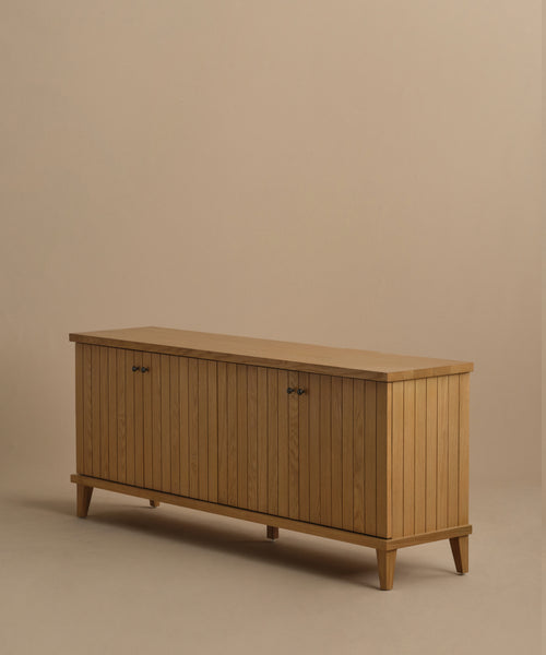The Coast Sideboard showcases a Mid-Century Modern style with vertical slats, warm oak finish, four small handles, and angled legs, displayed on a plain beige background.